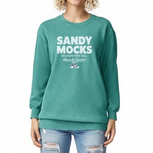 May include: Teal crew neck sweatshirt featuring the text "SANDY MOCKS" in white, with "MOCKUPS THAT SELL" and "Made for Creators" underneath. The sweatshirt is a solid teal colour and has long sleeves.