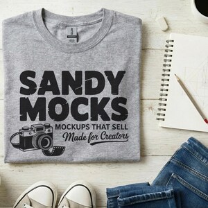 以下が含まれることがあります： 「SANDY MOCKS」の文字とカメラのグラフィックが入ったグレーのTシャツ。木製の表面に折りたたまれており、ノート、ペン、鉛筆、コーヒーカップ、ジーンズが近くにあります。スニーカーと小さな植物も見えます。