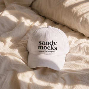 May include: A white baseball cap with the text "sandy mocks created for designers" in black. The cap is on a cream-colored, textured surface.
