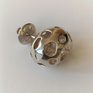 Puede incluir: Un pendiente plateado con un diseño único. El pendiente presenta un cuerpo hueco de forma ovalada con múltiples recortes circulares. El poste y el respaldo también son plateados, creando un aspecto cohesivo. El pendiente está diseñado para ser usado en la oreja.