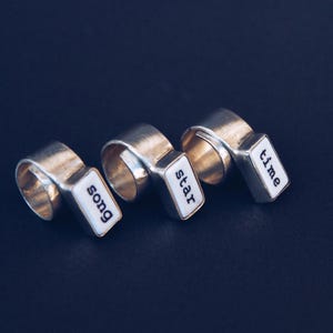 Puede incluir: Tres anillos de plata con incrustaciones rectangulares de esmalte blanco. Cada incrustación presenta una sola palabra en negro: "song", "star" y "time". Los anillos se muestran sobre una superficie azul oscuro.