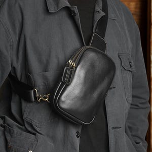 May include: A black leather crossbody sling bag with a smooth, rounded rectangular shape. The bag features two main zippered compartments with dark metal hardware, a wide adjustable black fabric strap, and a brass-toned metal swivel clip attachment.