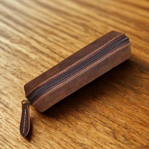 K&ouml;nnte beinhalten: Ein braunes Leder-Federm&auml;ppchen mit Rei&szlig;verschluss und kleinem Leder-Zuglasche. Das rechteckige Etui hat wei&szlig;e N&auml;hte an den Kanten. Das Federm&auml;ppchen ist aus einem Leder mit Used-Look gefertigt.