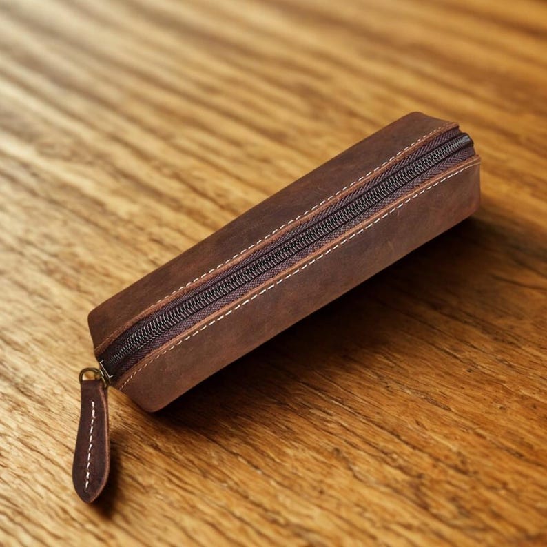 K&ouml;nnte beinhalten: Ein braunes Leder-Federm&auml;ppchen mit Rei&szlig;verschluss und kleinem Leder-Zuglasche. Das rechteckige Etui hat wei&szlig;e N&auml;hte an den Kanten. Das Federm&auml;ppchen ist aus einem Leder mit Used-Look gefertigt.