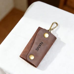 May include: A brown leather key holder with a gold-colored clasp and two gold-colored snap closures. The word "NAME" is printed vertically on the front. The key holder is made of a folded piece of leather.