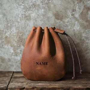 May include: A brown leather drawstring pouch featuring a smooth texture and a cinched top closure with matching leather cords. The word NAME is embossed in black capital letters on the front centre of the pouch, which has a rounded base.