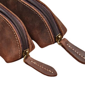 K&ouml;nnte beinhalten: Nahaufnahme von zwei braunen Lederbeuteln mit Messingrei&szlig;verschl&uuml;ssen und Ziern&auml;hten. Die Beutel sind aus Leder mit Used-Look gefertigt und die Rei&szlig;verschl&uuml;sse sind geschlossen. Die Beutel dienen wahrscheinlich zur Aufbewahrung kleiner Gegenst&auml;nde.