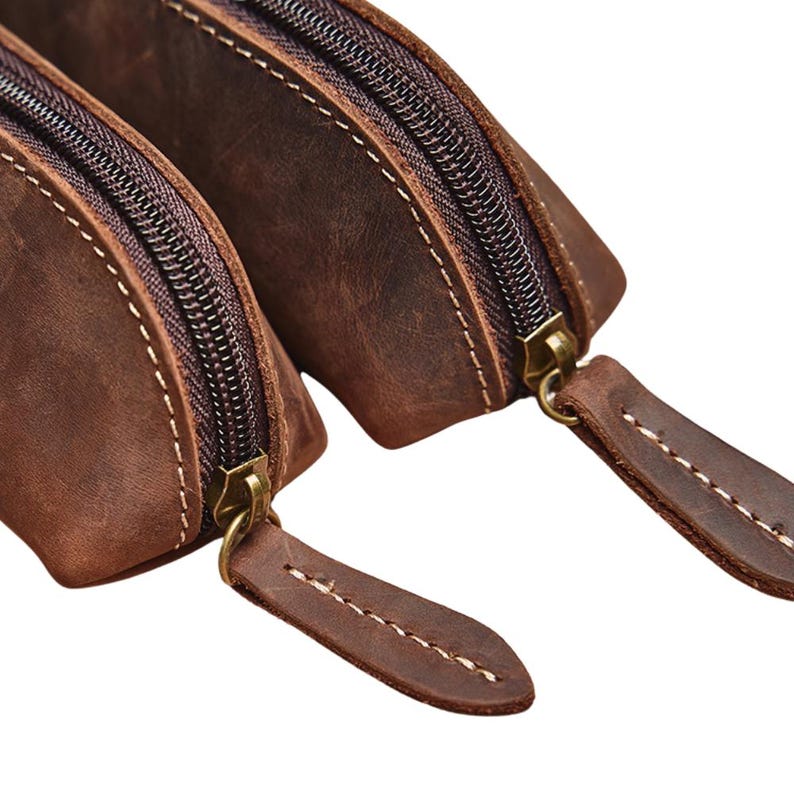 K&ouml;nnte beinhalten: Nahaufnahme von zwei braunen Lederbeuteln mit Messingrei&szlig;verschl&uuml;ssen und Ziern&auml;hten. Die Beutel sind aus Leder mit Used-Look gefertigt und die Rei&szlig;verschl&uuml;sse sind geschlossen. Die Beutel dienen wahrscheinlich zur Aufbewahrung kleiner Gegenst&auml;nde.