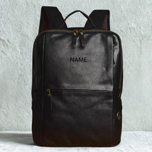 May include: Black leather backpack with a top handle, adjustable shoulder straps, and multiple zippered compartments. The word "NAME" is embossed on the front. Features brass-colored zippers and hardware. A stylish and functional bag for everyday use.