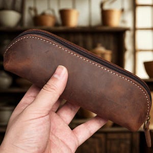 K&ouml;nnte beinhalten: Ein braunes Leder-Federm&auml;ppchen mit Rei&szlig;verschluss und wei&szlig;en N&auml;hten. Das Federm&auml;ppchen ist aus Leder im Used-Look gefertigt und hat einen kleinen Leder-Rei&szlig;verschluss.