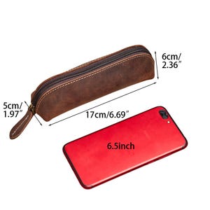 K&ouml;nnte beinhalten: Ein braunes Leder-Federm&auml;ppchen mit Rei&szlig;verschluss. Das Etui ist 17 cm lang, 5 cm breit und 6 cm hoch. Ein rotes Smartphone daneben misst 6,5 Zoll. Ein praktisches Accessoire f&uuml;r Stifte.