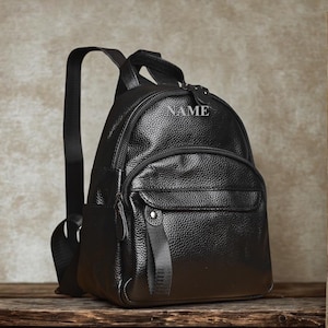 May include: Black leather backpack with a textured finish. Features a top handle, adjustable shoulder straps, and a front pocket. The word "NAME" is embossed on the top. The backpack has a zippered main compartment and a smaller front pocket.