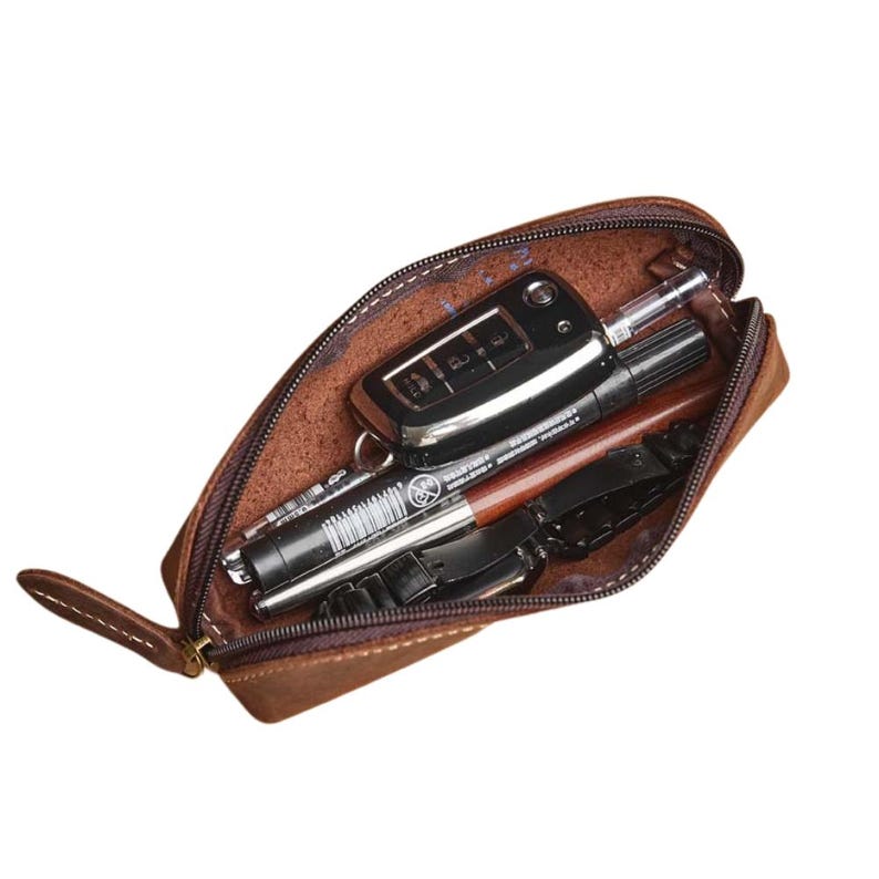 K&ouml;nnte beinhalten: Ein braunes Leder-Federm&auml;ppchen mit Rei&szlig;verschluss, das einen Autoschl&uuml;sselanh&auml;nger, einen Stift und andere kleine Gegenst&auml;nde enth&auml;lt. Das Etui hat eine Schlaufe an der Seite und ist ge&ouml;ffnet.