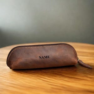 K&ouml;nnte beinhalten: Ein braunes Leder-Federm&auml;ppchen mit Rei&szlig;verschluss und kleinem Leder-Zugband. Das Wort "NAME" ist auf der Vorderseite aufgedruckt. Das Etui liegt auf einer Holzoberfl&auml;che.