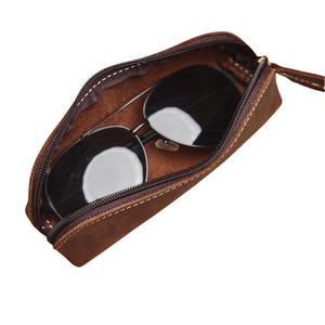 K&ouml;nnte beinhalten: Ein braunes Leder-Sonnenbrillenetui mit Rei&szlig;verschluss, das eine Sonnenbrille mit schwarzen Gl&auml;sern und silbernen Rahmen enth&auml;lt. Das Etui ist ge&ouml;ffnet und zeigt das Innenfutter und die N&auml;hte.
