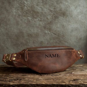 Könnte beinhalten: Eine braune Leder-Bauchtasche mit Reißverschluss und goldfarbener Schnalle. Das Wort "NAME" ist auf die Vorderseite der Tasche gedruckt. Die Tasche liegt auf einer Holzoberfläche.