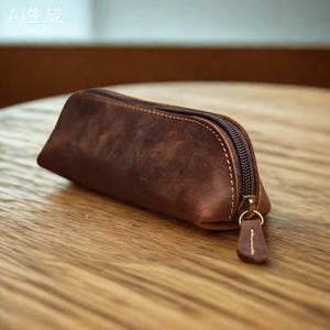 K&ouml;nnte beinhalten: Ein braunes Leder-Federm&auml;ppchen mit Rei&szlig;verschluss. Das Etui hat eine rechteckige Form mit abgerundeten Kanten und ist aus Leder. Der Rei&szlig;verschluss ist dunkelbraun, und der Zipper ist ein kleines St&uuml;ck Leder.