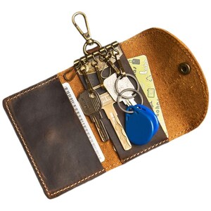 Könnte beinhalten: Ein braunes Schlüsseletui aus Leder mit Druckknopfverschluss, ausgestattet mit einem messingfarbenen Metallschlüsselhalter mit mehreren Haken, zwei Metallschlüsseln und einem blauen Kunststoff-Chip. Innen befinden sich Kartenfächer.