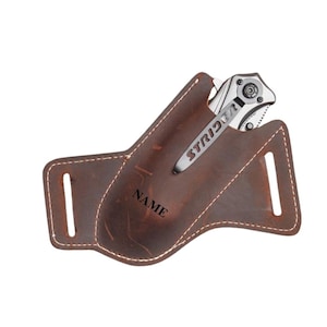 May include: A brown leather knife sheath with the word "NAME" embossed on the front. A silver folding knife with the word "STRIDER" on the clip is partially inserted into the sheath. The sheath has two vertical slots.