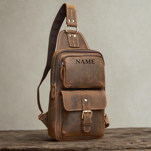 May include: A brown leather sling backpack featuring a single adjustable shoulder strap, a zippered top compartment with the word NAME embossed on the front, and a lower pocket secured with a metal buckle and a snap button closure.
