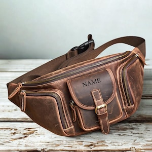 May include: A brown leather fanny pack featuring multiple zippered compartments, a front flap pocket with a buckle closure, and the word NAME engraved on the front. The bag includes an adjustable strap with a black plastic side-release buckle.