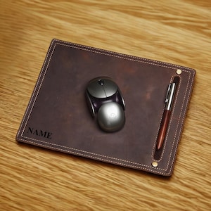 May include: A brown leather desk pad with a matching pen and a silver computer mouse. The pad has white stitching around the edges and the word "NAME" printed in the corner. The pen rests in a built-in holder.