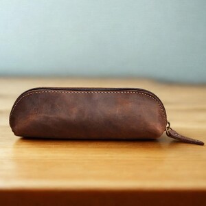 K&ouml;nnte beinhalten: Ein braunes Leder-Federm&auml;ppchen mit Rei&szlig;verschluss und kleinem Zugband. Das ovale Etui hat sichtbare N&auml;hte entlang der N&auml;hte. Es liegt auf einer Holzoberfl&auml;che.