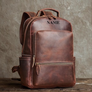 May include: A dark brown leather backpack featuring a top handle, adjustable shoulder straps, and a front zippered pocket. The word NAME is embossed in black on the upper front section. The bag includes side pockets and metal zipper pulls.
