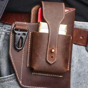 May include: A brown leather belt holster featuring a vertical pocket with a snap-button flap closure, a side-mounted black metal carabiner clip, and a dedicated slot containing a red smartphone and a rectangular silver-toned object.