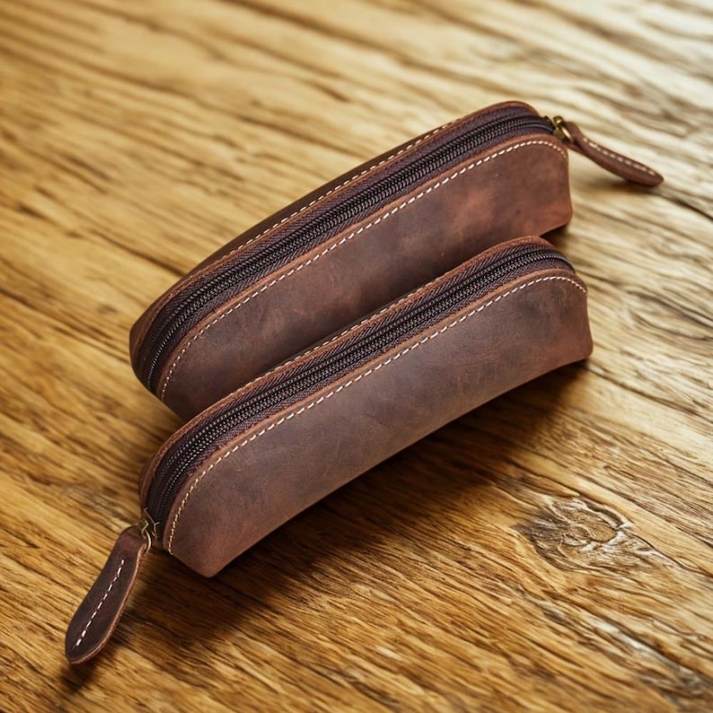 K&ouml;nnte beinhalten: Zwei braune Leder-Federm&auml;ppchen mit Rei&szlig;verschluss und wei&szlig;en N&auml;hten. Die Etuis sind unterschiedlich gro&szlig; und haben einen Zipper am Rei&szlig;verschluss. Die Etuis liegen auf einer Holzoberfl&auml;che.