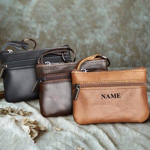 May include: Three small leather coin purses in shades of black, brown, and tan. Each purse has a zipper closure and a small handle. The tan purse has the word "NAME" engraved on it.