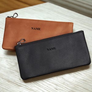 May include: Two leather pouches, one in a rich brown and the other in black, are displayed. Each pouch has a zipper closure, a silver-tone clasp, and the word "NAME" printed on the front. The pouches are rectangular and appear to be made of textured leather.
