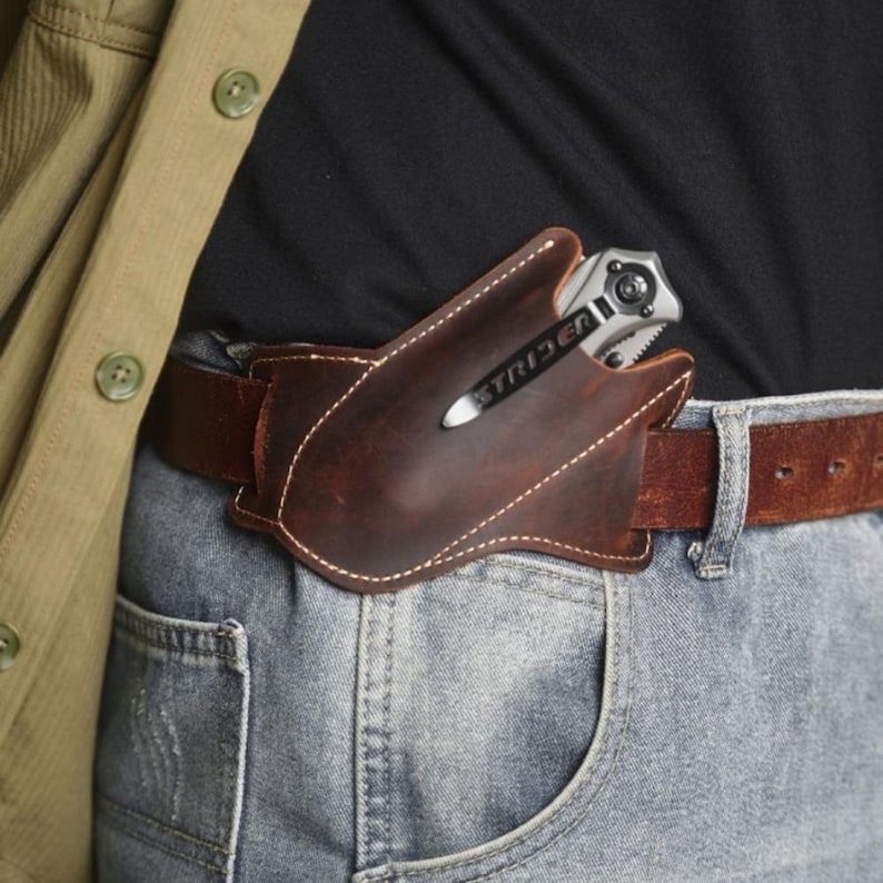Peut inclure: Un &eacute;tui &agrave; couteau en cuir marron avec des coutures blanches, port&eacute; sur une ceinture marron. Un couteau pliant argent&eacute; avec le mot "STRIDER" est partiellement visible dans l'&eacute;tui. La personne porte un jean bleu et une veste verte.