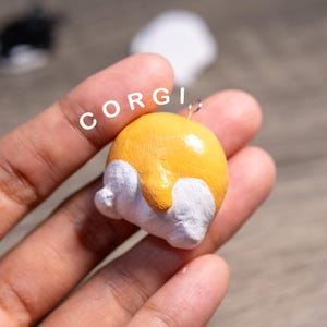 May include: A handmade corgi butt charm in yellow and white. The charm is round and has a small metal loop at the top. The word "CORGI" is written in white letters above the charm.