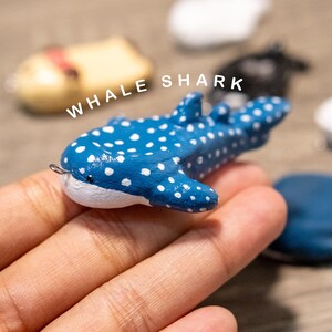 Könnte beinhalten: Blaue Walhai-Figur mit weißen Punkten und weißem Bauch. Der Text "WHALE SHARK" befindet sich über der Figur. Die Figur wird in einer Hand gehalten.