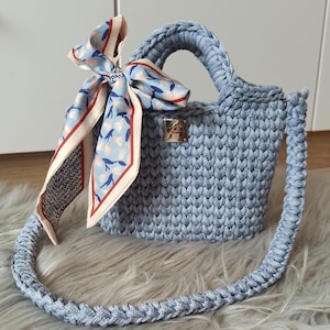 May include: A light blue crocheted handbag with a matching braided strap and handle. A patterned scarf in shades of blue, red, and cream is tied to the handle. The bag features a small, rectangular metal emblem.