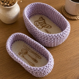 May include: Two oval-shaped baskets made of light purple crocheted yarn with wooden bases. The larger basket features a laser-engraved croissant design, while the smaller basket displays an engraved illustration of a fork, spoon, and knife.