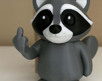 Puede incluir: Figura de mapache gris impresa en 3D con marcas faciales en blanco y negro. El mapache está mostrando el dedo medio. Una regla de madera está en el fondo, midiendo aproximadamente 18 cm de altura.