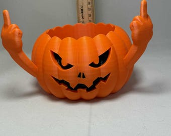 Puede incluir: Una calabaza naranja impresa en 3D con una cara tallada y brazos levantados, cada mano mostrando el dedo medio. La calabaza tiene un borde dentado y cuencas de ojos negras. Un artículo decorativo con temática de Halloween.