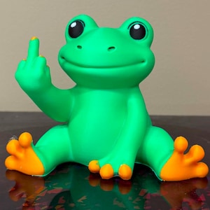 May include: A bright green frog figurine with large black eyes and orange-tipped toes. The frog is in a seated position with one arm raised, extending its middle finger in a gesture. The surface of the frog has a smooth, matte finish.