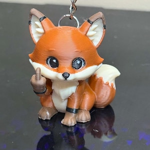 May include: A 3D printed fox keychain featuring a rust-orange and cream-colored fox figure with large black eyes. The fox is depicted in a seated position, holding up its right paw with the middle finger extended in a gesture, attached to a metal chain.