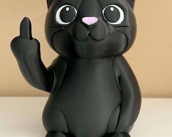 Puede incluir: Figura de gato negro con ojos blancos, nariz rosa y dedo corazón levantado. El gato está sentado y tiene un acabado liso y mate. Un artículo decorativo humorístico y único.