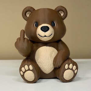 May include: A brown 3D printed bear figurine with a cream-colored belly, muzzle, and paw pads. The bear has black eyes and a black nose, and its right arm is raised with the middle finger extended in a gesture. The surface has visible print lines.