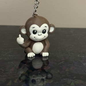 May include: A brown monkey keychain with white accents, giving the middle finger. The monkey has a white belly and ears, with a silver keychain ring attached. The monkey is sitting down and has a smiling face.