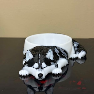 May include: A small, white, circular bowl integrated into a 3D-printed figurine of a resting Siberian Husky dog. The dog features black and white fur patterns, pointed ears, and is lying down with its head resting on its front paws.