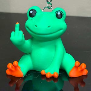 May include: A bright green frog keychain with orange feet and black eyes. The frog is in a seated position and is holding up its right middle finger. A silver metal chain and ring are attached to the top of the frog's head for hanging purposes.