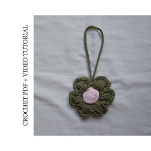 Può includere: Un fiore all'uncinetto fatto a mano con un centro rosa e petali verde oliva, sospeso da un anello abbinato. Il testo "CROCHET PDF + VIDEO TUTORIAL" è stampato verticalmente a sinistra.