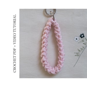 以下が含まれることがあります： シルバーカラーの金属リングが付いた、薄いピンク色の編み物キーホルダー。ストラップは太い編み糸で作られています。左側のテキストは「CROCHET PDF + VIDEO TUTORIAL」と書かれています。
