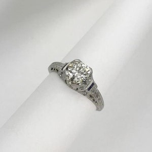 May include: A silver engagement ring featuring a round, clear diamond centre stone and two small blue sapphire accent stones.