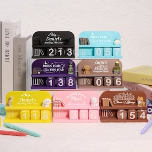 May include: A selection of book-themed desk organisers in various colours. Each has a shelf with a name, decorative items, and numbered blocks for tracking books read. Colours include black, blue, purple, brown, yellow, and pink.